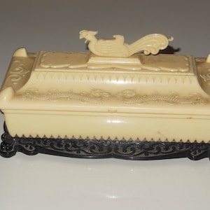 May include: A cream-colored decorative box with a black base. The box has an ornate design and a bird-shaped lid.