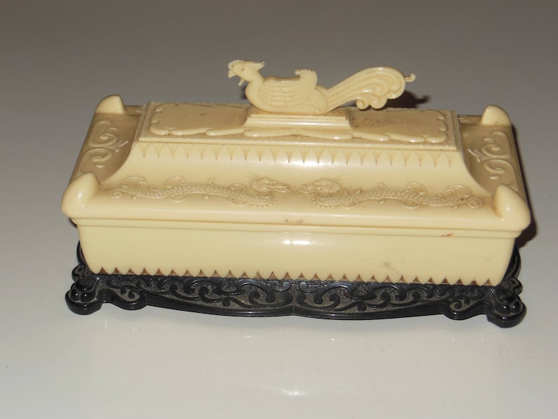May include: A cream-colored decorative box with a black base. The box has an ornate design and a bird-shaped lid.