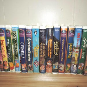 Pu&ograve; includere: Una collezione di videocassette VHS Disney, con titoli come "Il libro della giungla", "Aladdin", "Pocahontas", "Cenerentola" e "La bella e la bestia". Le cassette sono disposte in fila, con dorsi colorati e titoli di film.