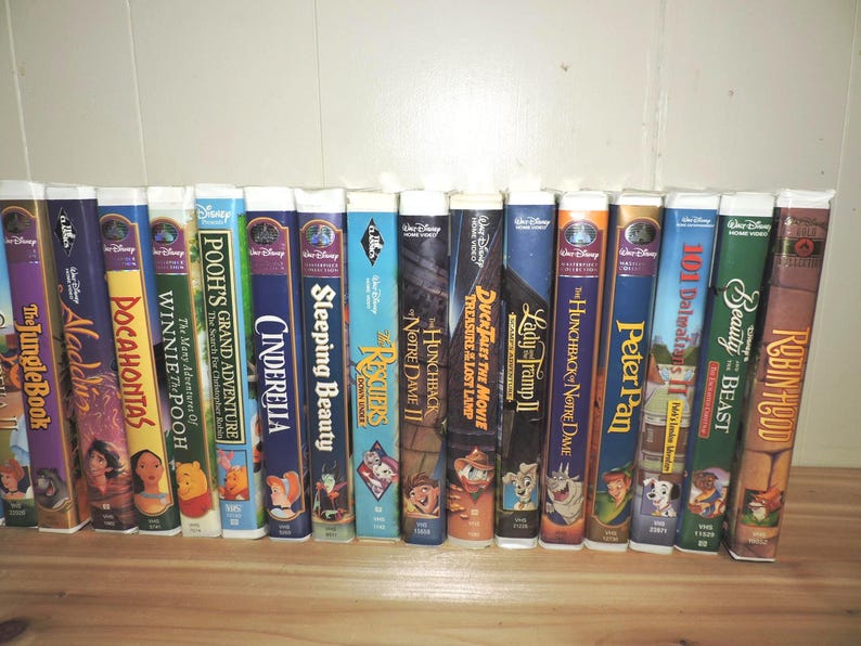 Pu&ograve; includere: Una collezione di videocassette VHS Disney, con titoli come "Il libro della giungla", "Aladdin", "Pocahontas", "Cenerentola" e "La bella e la bestia". Le cassette sono disposte in fila, con dorsi colorati e titoli di film.