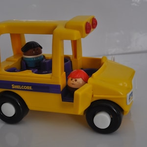 Può includere: Un autobus scolastico giocattolo giallo con una striscia viola e la parola "Shelcore" stampata sul lato. L'autobus ha due finestre e due luci rosse sul tetto. Ci sono due figure all'interno dell'autobus, una con la pelle marrone e una con i capelli rossi.