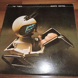Könnte beinhalten: Ein Vintage-Vinyl-Plattencover mit einem futuristisch aussehenden Roboter, der ein Baby in einer schwarz-weißen Babytrage hält. Das Albumcover trägt den Titel "...THE TUBES... REMOTE CONTROL..."
