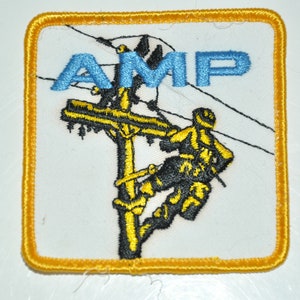 May include: A yellow embroidered patch with a white background. The patch features a lineman climbing a utility pole with the word "AMP" in blue lettering above the pole.