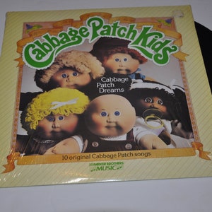 Puede incluir: Una portada de álbum de vinilo con cinco muñecas Cabbage Patch Kids. La portada es amarilla con un título verde que dice "Cabbage Patch Kids". El título está rodeado de una cinta amarilla. El título del álbum es "Cabbage Patch Dreams". La portada del álbum también incluye el texto "10 canciones originales de Cabbage Patch" y "Rainbow Brothers Music".