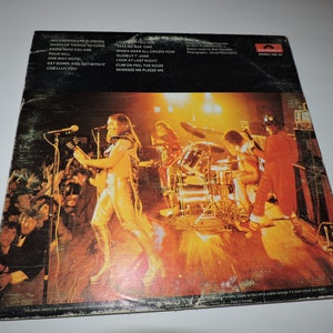 May include: A vintage vinyl record album cover featuring a live performance by a rock band. The band is on stage with a drummer, guitarist, and singer. The album cover is yellowed and has a worn look. The album title is "Wildfire" and the band name is "The Sweet".
