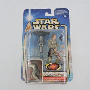 Op de afbeelding: Een Star Wars actiefiguur van Luke Skywalker in een plastic verpakking. De figuur draagt een bruin en beige outfit en houdt een lichtzwaard vast. De verpakking heeft het Star Wars-logo en de tekst "The Empire Strikes Back".