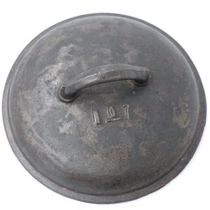 Puede incluir: Una tapa redonda de hierro fundido gris oscuro con un asa curvada. La tapa tiene el número "107" estampado en la superficie. El metal tiene un aspecto texturizado y envejecido, lo que sugiere que es vintage.