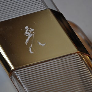 May include: A clear glass bottle with a gold-colored cap. The cap has a raised design of a man in a hat and a walking stick.