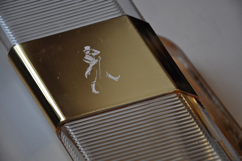 May include: A clear glass bottle with a gold-colored cap. The cap has a raised design of a man in a hat and a walking stick.