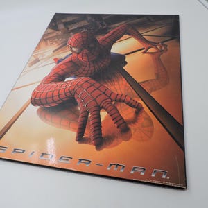 Op de afbeelding: Een promotieposter met Spider-Man in een dynamische pose. De superheld is afgebeeld in zijn iconische rood-blauwe pak, met een gedetailleerd webpatroon. De poster heeft de woorden "SPIDER-MAN" in zilveren letters.