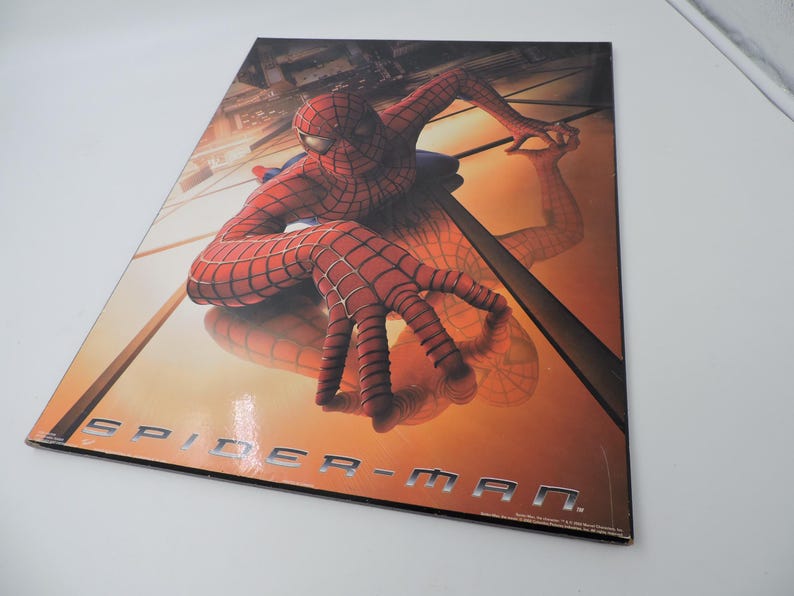 Op de afbeelding: Een promotieposter met Spider-Man in een dynamische pose. De superheld is afgebeeld in zijn iconische rood-blauwe pak, met een gedetailleerd webpatroon. De poster heeft de woorden "SPIDER-MAN" in zilveren letters.