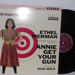 Peut inclure: Une pochette d'album de disque vinyle vintage mettant en vedette Ethel Merman dans la production de Rodgers & Hammerstein d'Annie Get Your Gun. La pochette présente une cible rouge et blanche avec une femme tenant un fusil dans une tenue brune et frangée.