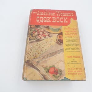 May include: Vintage cookbook titled "The American Woman's Cook Book" with a red and yellow cover. The cover features images of prepared food and text. The book is worn, showing signs of age and use. It includes recipes and household hints.
