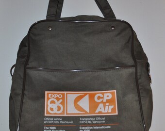 Pode incluir: Uma bolsa de lona verde com guarnição marrom e um logotipo da CP Air, a companhia aérea oficial da Expo 86 em Vancouver. O logotipo apresenta uma seta estilizada apontando para a esquerda e o texto "CP Air". A bolsa também inclui detalhes sobre a Exposição Mundial de 1986 em Vancouver, Colúmbia Britânica, Canadá.