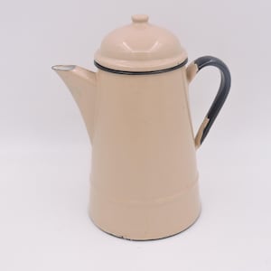 Könnte beinhalten: Eine Vintage-Kaffeekanne aus beigefarbenem Emaille mit Deckel und schwarzem Griff. Der Ausguss befindet sich auf der linken Seite, und die Kanne hat eine schwarze Umrandung um den Deckel und die Oberseite der Kanne. Die Kanne steht auf weißem Hintergrund.