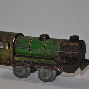 May include: A vintage toy train engine made of metal with a green and black paint scheme. The engine has a small cab, a large boiler, and four wheels. The toy is showing signs of wear and tear, with some rust and scratches.