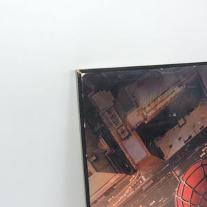 Op de afbeelding: Een ingelijste print met een stadsgezicht met een rood en zwart Spider-Man-masker op de voorgrond. Het kunstwerk toont een nachtelijk uitzicht op gebouwen en straten, met de nadruk op het iconische masker van de superheld.
