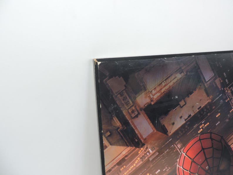 Op de afbeelding: Een ingelijste print met een stadsgezicht met een rood en zwart Spider-Man-masker op de voorgrond. Het kunstwerk toont een nachtelijk uitzicht op gebouwen en straten, met de nadruk op het iconische masker van de superheld.