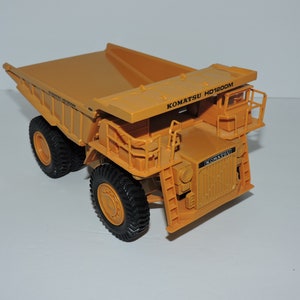 May include: A yellow toy dump truck with the text "KOMATSU HD1200M" on the side. The truck has a large bed and four wheels.