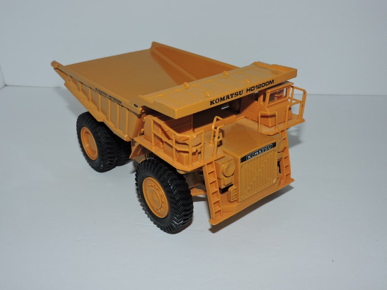 May include: A yellow toy dump truck with the text "KOMATSU HD1200M" on the side. The truck has a large bed and four wheels.