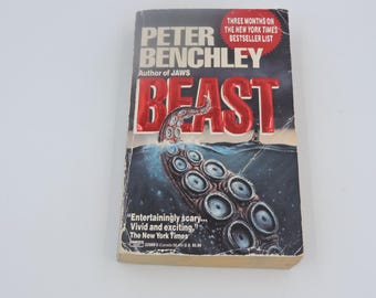 Op de afbeelding: Een vintage paperback boek getiteld "BEAST" van Peter Benchley, auteur van "JAWS". De cover toont een rode titel met een illustratie van een octopus tentakel die uit de oceaan komt. Het boek is een New York Times bestseller.