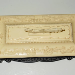 May include: A white ceramic trinket box with a black base. The box is decorated with a raised design of dragons and a bird. The lid of the box has a raised design of a bird.