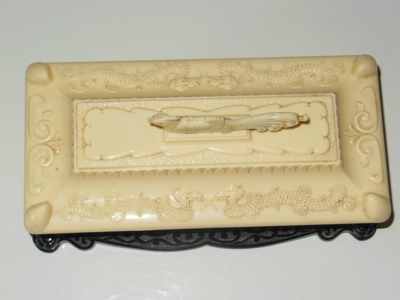 May include: A white ceramic trinket box with a black base. The box is decorated with a raised design of dragons and a bird. The lid of the box has a raised design of a bird.