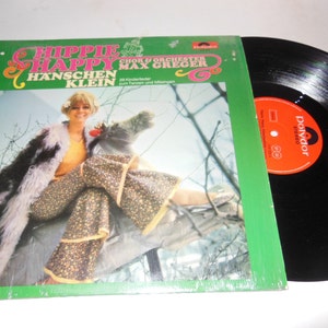 May include: A vinyl record album cover featuring a woman in a furry coat and floral trousers sitting on a tree branch. The album title is "Hippie & Happy" by Hanschen Klein, with a choir and orchestra led by Max Greger. The record label is Polydor.
