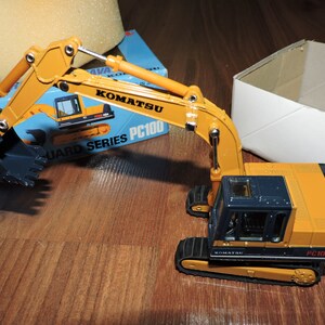 May include: A yellow and blue toy excavator with a black bucket. The excavator is labeled "Komatsu PC100 Guard Series".
