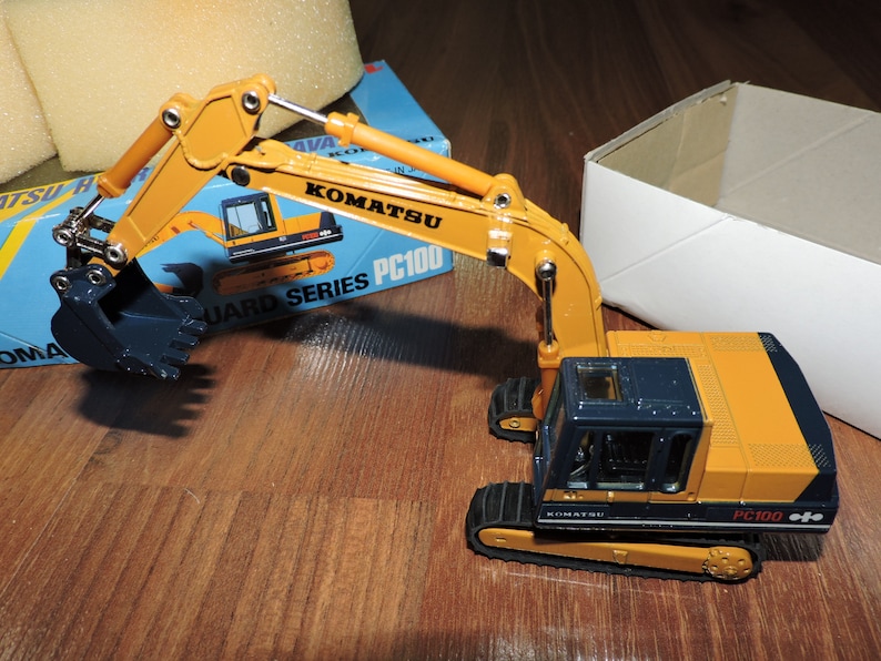 May include: A yellow and blue toy excavator with a black bucket. The excavator is labeled "Komatsu PC100 Guard Series".