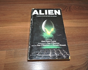 May include: A paperback book cover for the novel "Alien" by Alan Dean Foster. The cover features a black and green alien creature with a yellow glow. The text "In space no one can hear you scream." is printed on the cover.