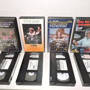 May include: Six VHS tapes of drum master classes and a concert. The tapes feature Carmine Appice, Steve Gadd, Tommy Aldridge, and Buddy Rich. The tapes are black with white labels and are arranged in two rows.