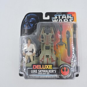 Op de afbeelding: Deluxe Luke Skywalker's Desert Sport Skiff actiefiguur, verpakt in een transparante plastic hoes. De verpakking toont het Star Wars-logo, een Darth Vader-afbeelding en het Rebel Alliance-symbool. Inclusief een Luke Skywalker-figuur.
