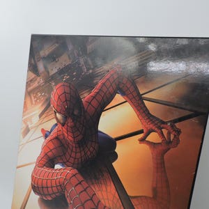 Op de afbeelding: Een ingelijste afbeelding met Spider-Man in zijn iconische rood-blauwe pak. De superheld wordt afgebeeld in een dynamische pose, zich vastklampend aan een reflecterend oppervlak met een stadsgezicht op de achtergrond. Het kunstwerk toont levendige kleuren en gedetailleerde texturen.