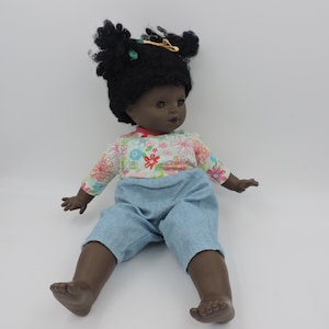 Może przedstawiać: Ciemnoskóra lalka z czarnymi kręconymi włosami, ubrana w koszulę z długim rękawem w kwiatowy wzór i jasnoniebieskie spodnie. Lalka ma brązowe oczy i siedzi z wyciągniętymi nogami. Złota spinka do włosów jest we włosach lalki.