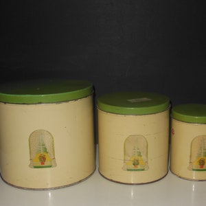 May include: Set of three vintage kitchen canisters with green lids and a cream base. Each canister features a printed design of a window with a potted plant inside.