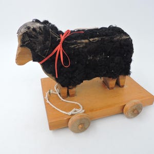 Peut inclure: Un mouton jouet à tirer vintage. Le mouton a un corps noir texturé, une tête en bois et est orné d'un ruban rouge. Il repose sur une base en bois avec quatre roues en bois et une ficelle blanche.