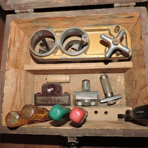Op de afbeelding: Een houten kist met een verscheidenheid aan gereedschappen en instrumenten erin. De kist is open en de gereedschappen zijn zichtbaar. De gereedschappen omvatten een metalen apparaat met een handvat, een houten pen, een metalen klem, een metalen pons, een metalen staaf en een schroevendraaier.