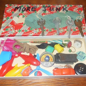 Puede incluir: Una caja abierta llena de varios juguetes pequeños y baratijas, incluyendo un coche de juguete rojo y azul, un avión de juguete rojo y amarillo, una cuerda marrón, un juego de juguete azul y blanco y un juguete rojo y blanco con las palabras "Put Balls in Hole" en él. La caja está decorada con estrellas rojas y verdes y las palabras "More Junk" están escritas con marcador negro en la parte superior de la caja.