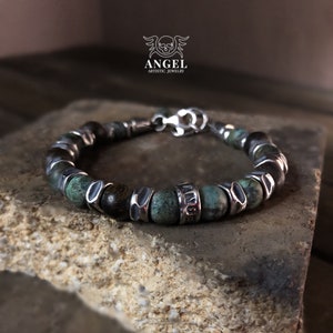 May include: A beaded bracelet featuring turquoise and dark brown beads, accented with silver-toned metal spacers and a clasp. The bracelet is displayed on a textured stone surface. The word "ANGEL" is visible on the bracelet.