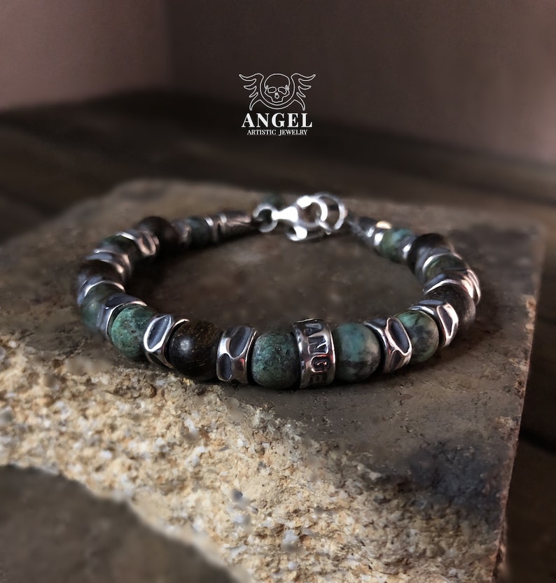 May include: A beaded bracelet featuring turquoise and dark brown beads, accented with silver-toned metal spacers and a clasp. The bracelet is displayed on a textured stone surface. The word "ANGEL" is visible on the bracelet.