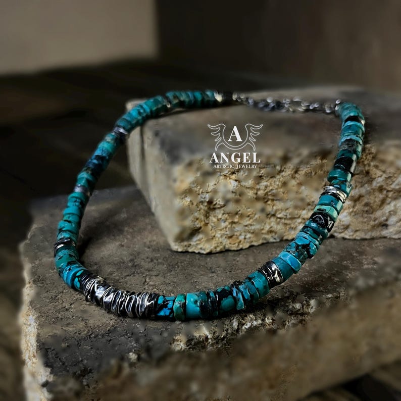 May include: A turquoise and black beaded necklace with silver accents. The necklace is made of small, round turquoise beads interspersed with black and silver elements. The necklace is displayed on a stone surface. The logo "ANGEL ARTISTIC JEWELRY" is visible.