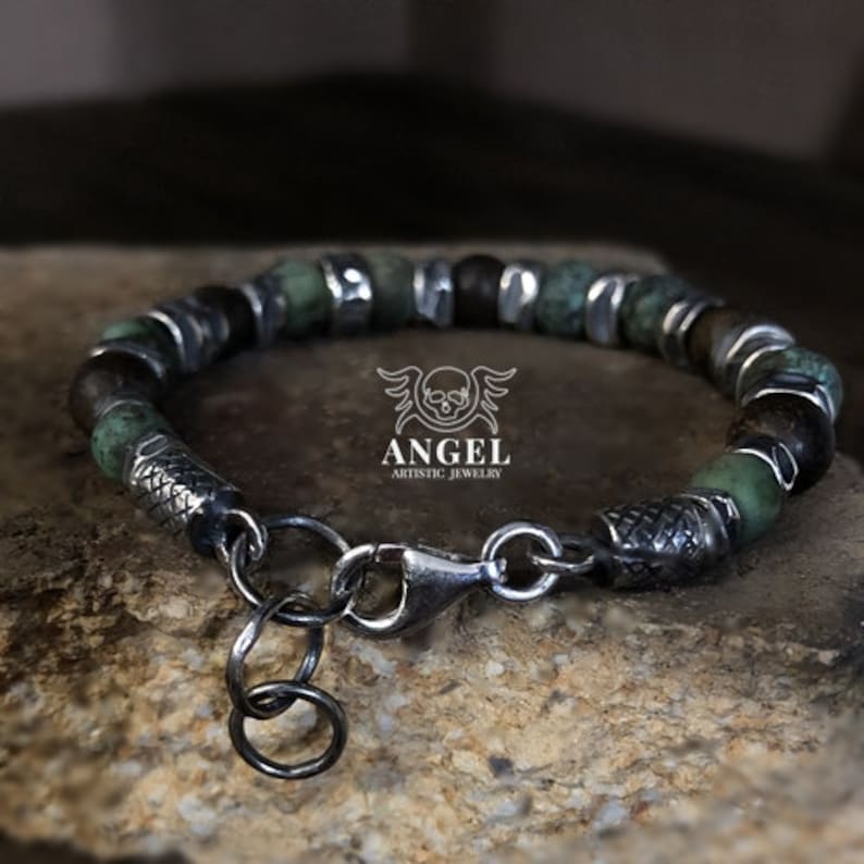 May include: A beaded bracelet featuring green and brown stone beads interspersed with silver-toned metal accents. The bracelet has a lobster clasp closure and a small charm. The brand name "ANGEL ARTISTIC JEWELRY" is visible.
