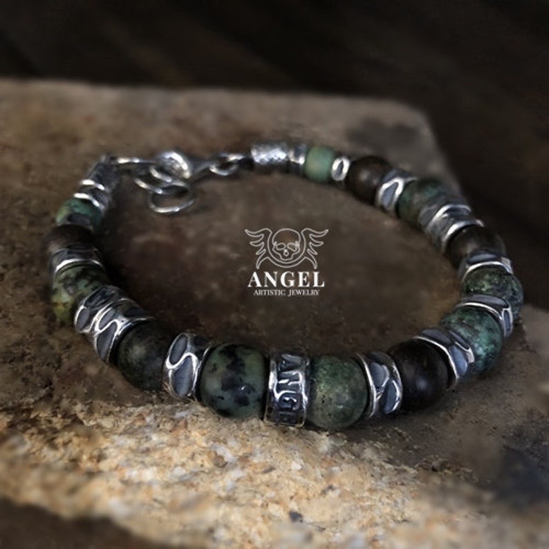 May include: A beaded bracelet featuring green and brown stone beads interspersed with silver-toned metal accents. The bracelet has a lobster clasp closure and the word "ANGEL" is engraved on one of the metal beads. The logo "ANGEL ARTISTIC JEWELRY" is also visible.