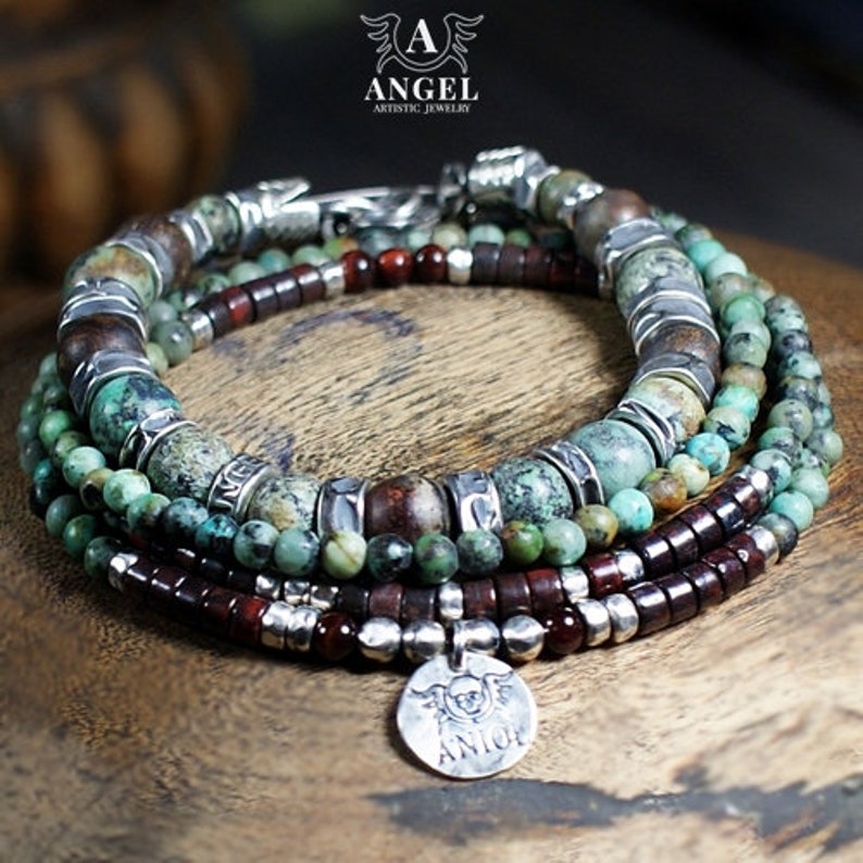 May include: A multi-strand bracelet featuring turquoise, brown, and silver beads. The bracelet includes a silver charm with an engraved design. The jewelry is displayed on a wooden surface.