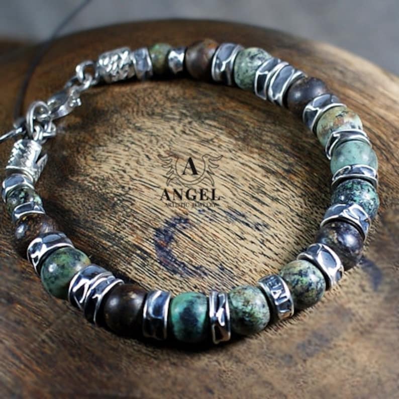 May include: A beaded bracelet featuring a mix of green, brown, and silver beads. The bracelet has a chain closure and is displayed on a wooden surface. The text "ANGEL ARTISTIC JEWELRY" is visible.