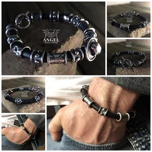 May include: A black leather bracelet with silver-toned metal beads and accents. The bracelet features a toggle clasp and various decorative elements, including patterned and textured beads. The brand logo "ANGEL ARTISTIC JEWELRY" is visible.