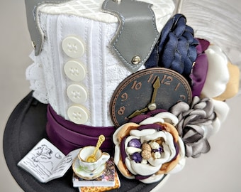 May include: A white top hat with a gray leather band, a clock face, and a variety of fabric flowers in shades of purple, white, and gray. The hat also features a purple ribbon and a small book with a drawing of a rabbit.