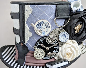 May include: A black top hat with a blue and white quilted fabric top, decorated with lace, a teacup and saucer, a clock face, and a small book. The hat has a black and white striped band around the bottom.