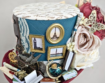 May include: A teal blue top hat with a white lace trim and a variety of embellishments, including a miniature Eiffel Tower, a compass, and a globe. The hat is decorated with a floral arrangement of pink and white roses and a butterfly.